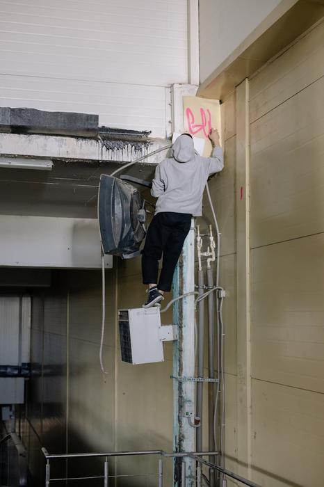 A street artist in a hoodie spray paints graffiti on a tall industrial wall.