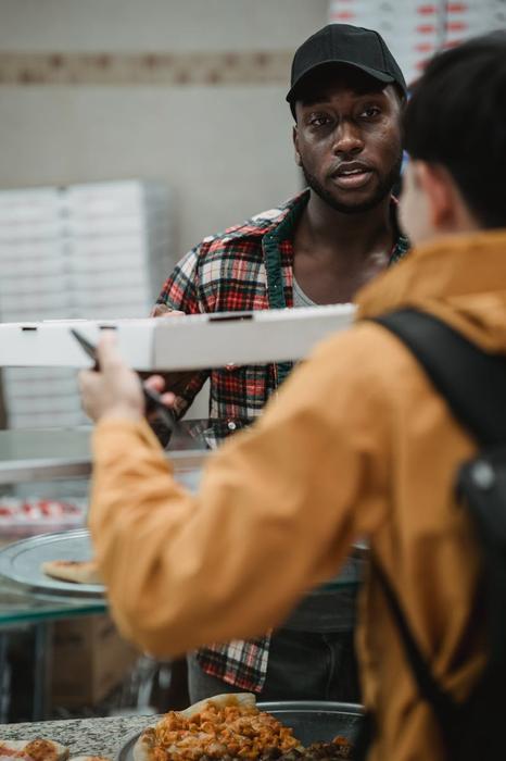 A man in a plaid shirt serves pizza to a customer at a restaurant counter.